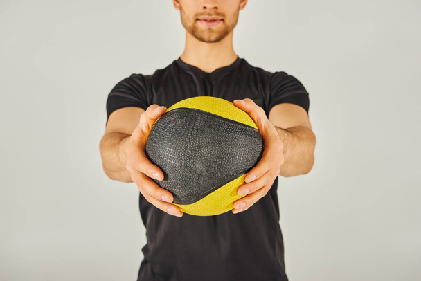 Un joven deportista en ropa activa sostiene una bola amarilla y negra en un estudio con un fondo gris. - Foto, imagen