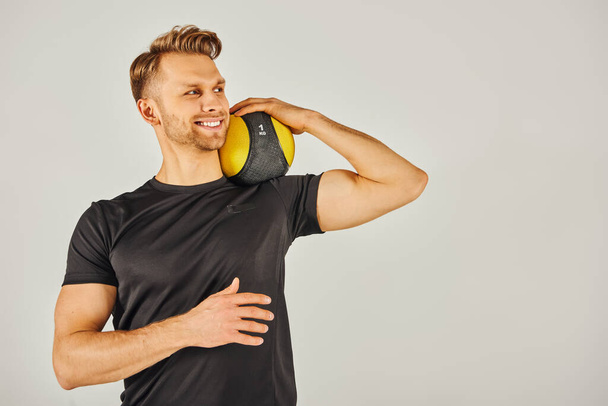 Un joven deportista en ropa activa sosteniendo una pelota amarilla, mostrando equilibrio y enfoque en un estudio con un fondo gris. - Foto, Imagen