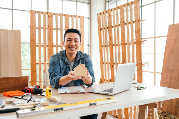 Portrait of a smile Asian male father sitting and working with a laptop on the table with the instrument of measure and working tools on the table at home woodwork studio. - 写真・画像