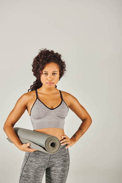 Sportiva afroamericana riccia in abbigliamento attivo con in mano un tappetino da yoga in uno studio con sfondo grigio. - Foto, immagini