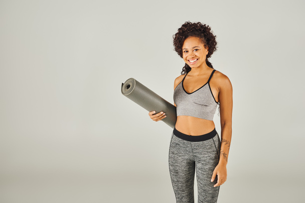 Eine lockige afroamerikanische Sportlerin in Activwear hält in einem Studio mit grauem Hintergrund eine Yogamatte in der Hand. - Foto, Bild