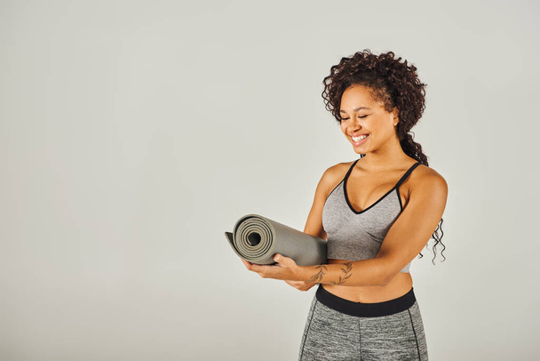 Eine lockige afroamerikanische Sportlerin in sportlicher Kleidung hält in einem Studio vor grauem Hintergrund eine Yogamatte in der Hand. - Foto, Bild