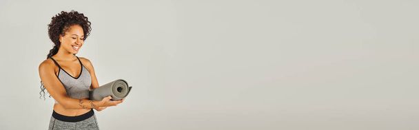 Afroameričanka ve sportovním oblečení držení jóga podložka a cvičení ve studiu s šedým pozadím. - Fotografie, Obrázek