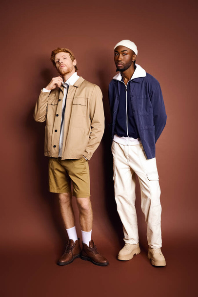 Два мультикультурных мужчины со стильной одеждой стоят рядом друг с другом перед коричневой стеной. - Фото, изображение