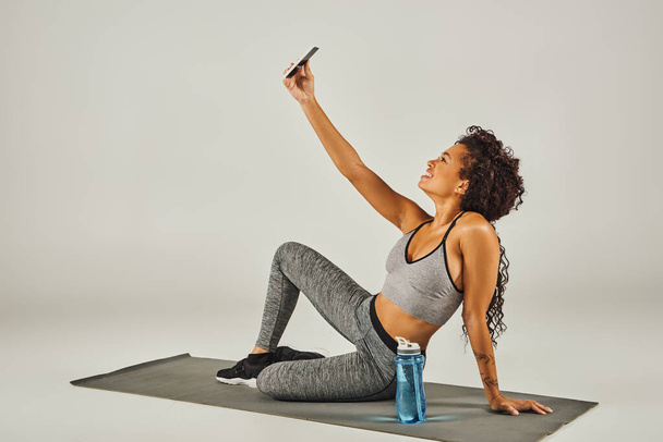 Une sportive afro-américaine frisée prend un selfie assis sur un tapis de yoga dans un studio avec un fond gris. - Photo, image