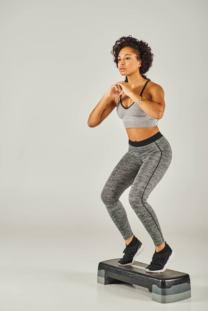 Kręcone Afroamerykanka sportowa w aktywnym nosić energicznie kroki na kroczku w studio z szarym tle. - Zdjęcie, obraz