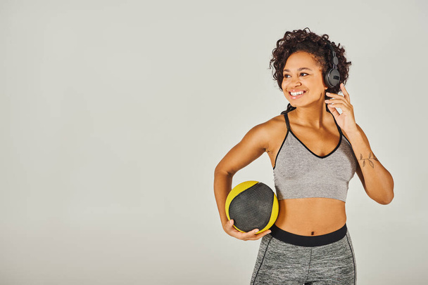 Kiharatukkainen afroamerikkalainen nainen urheiluvaatteissa, kuulokkeet päässä, pallo kädessä, - Valokuva, kuva