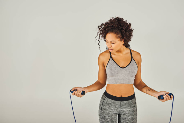 Una deportista afroamericana rizada en ropa activa sosteniendo una cuerda saltando frente a un fondo gris. - Foto, imagen