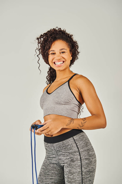 Eine lockige afroamerikanische Sportlerin in Activwear lächelt, während sie in einem Studio vor grauem Hintergrund ein Springseil hält. - Foto, Bild