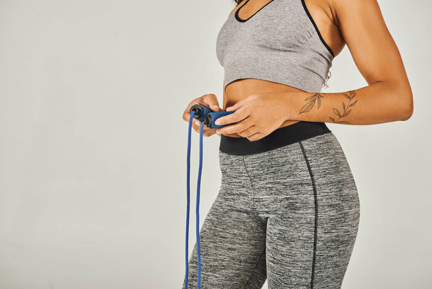 Афроамериканская спортсменка в активной одежде энергично держит скакалку в студии на сером фоне. - Фото, изображение