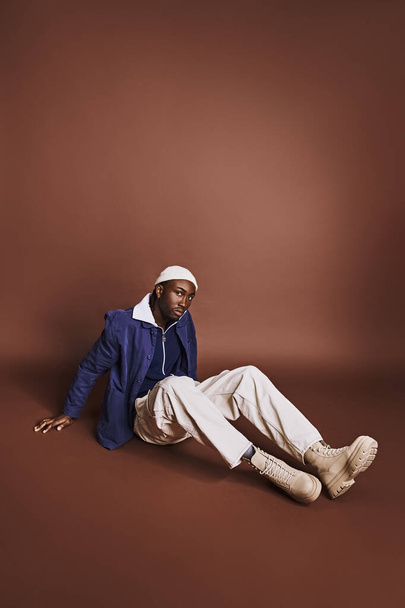 Fešák Afroameričan sedí na zemi v modré bundě a bílých kalhotách. - Fotografie, Obrázek