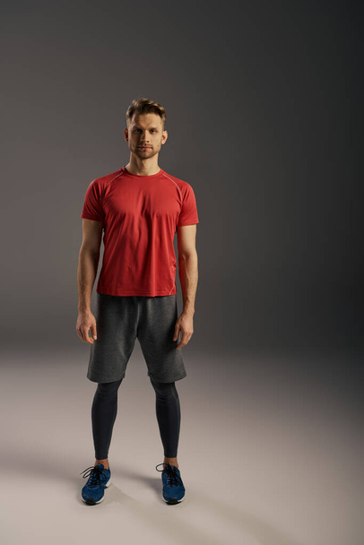 Un hombre con una camiseta roja y pantalones cortos grises asoma con confianza una pose frente a un fondo gris sereno. - Foto, imagen