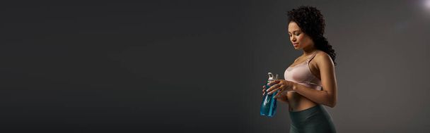 Кудрявая афроамериканская спортсменка держит бутылку воды во время тренировки в студии на сером фоне. - Фото, изображение