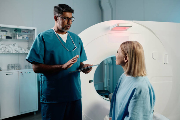 Elinde dijital tablet bulunan çift ırklı radyograf MRI odasında yetişkin beyaz kadın hastalarla çalışıyor. - Fotoğraf, Görsel