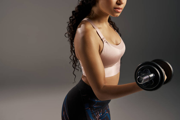 Egy göndör hajú afro-amerikai sportos nő aktív ruhában súlyzókkal dolgozik szürke stúdió háttérben.. - Fotó, kép