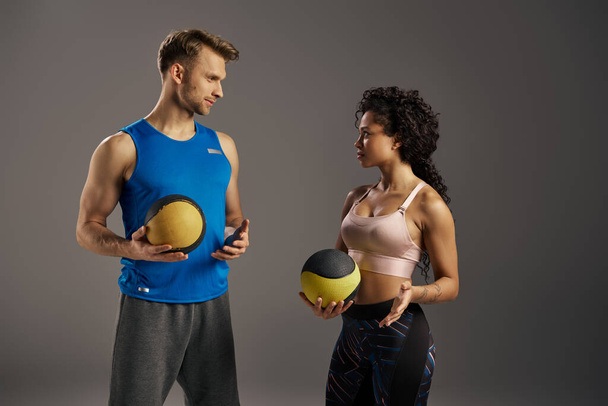 Młoda wielokulturowa i wysportowana para w aktywnym ubraniu stojąca obok siebie, trzymająca piłki treningowe w studio. - Zdjęcie, obraz