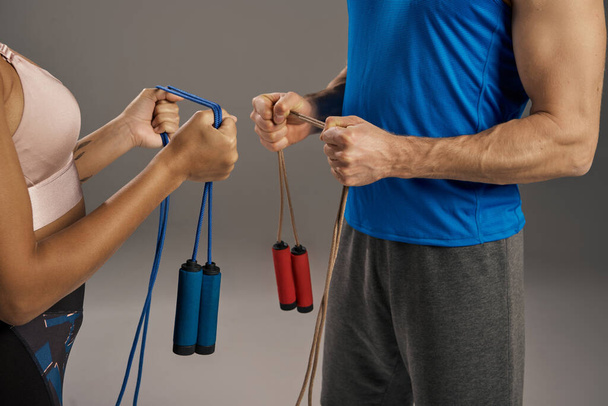 Jovem casal multicultural em equipamento de treino, segurando cordas de salto, pronto para iniciar sua rotina de fitness em um fundo de estúdio cinza. - Foto, Imagem