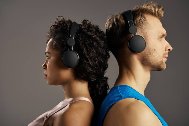 Młoda, wielokulturowa para w stroju aktywnym, zanurzona w muzyce, nosząca słuchawki podczas treningu w studio na szarym tle. - Zdjęcie, obraz