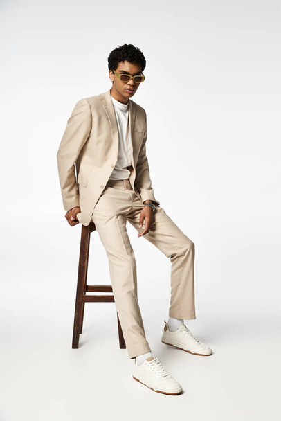 Przystojny Afroamerykanin w brązowym garniturze siedzi na stołku w stylowych okularach przeciwsłonecznych. - Zdjęcie, obraz