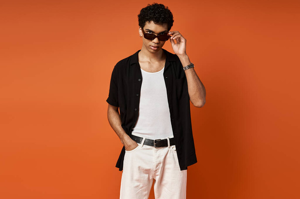 Όμορφος άντρας με γυαλιά ηλίου σε μαύρο πουκάμισο και λευκό παντελόνι σε στάση απέναντι σε έντονο πορτοκαλί φόντο. - Φωτογραφία, εικόνα
