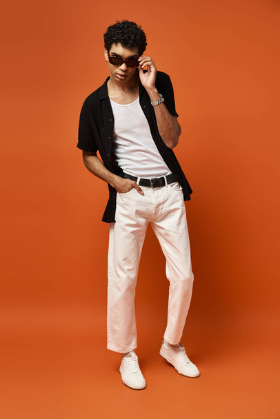 Handsome man in white pants and black shirt poses on vibrant orange backdrop. - Photo, Image