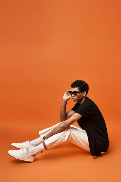 Przystojny Afroamerykanin siedzący na ziemi w okularach przeciwsłonecznych i białych butach. - Zdjęcie, obraz