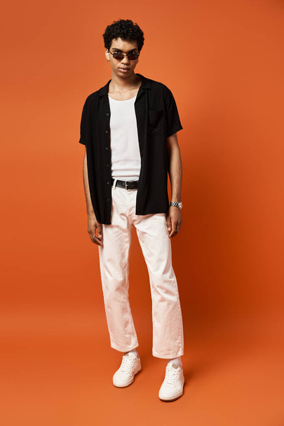 Αφροαμερικάνος με λευκό παντελόνι και μαύρο πουκάμισο στέκεται σε έντονο πορτοκαλί φόντο. - Φωτογραφία, εικόνα