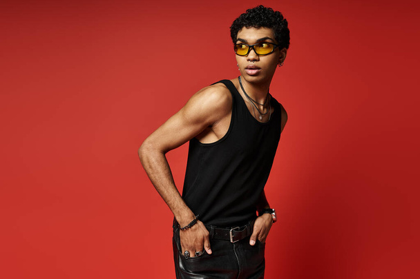 Przystojny Afroamerykanin pozujący w stylowych okularach przeciwsłonecznych, czarnej koszulce i skórzanych spodniach. - Zdjęcie, obraz