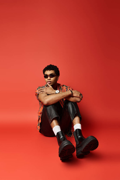 Αφροαμερικάνος με γυαλιά ηλίου κάθεται σε κόκκινο φόντο με μαύρες μπότες. - Φωτογραφία, εικόνα