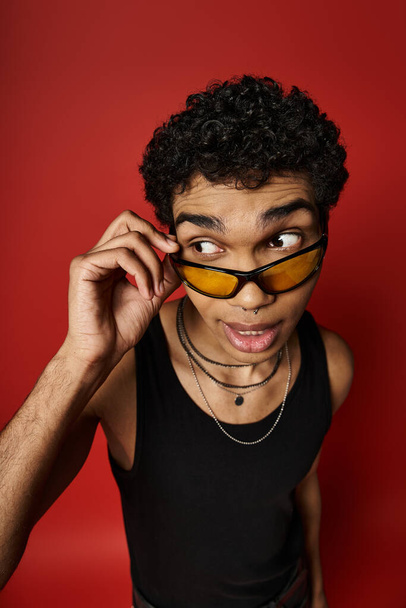 Een jongeman straalt coole vibraties uit in een zonnebril tegen een gewaagde rode achtergrond. - Foto, afbeelding