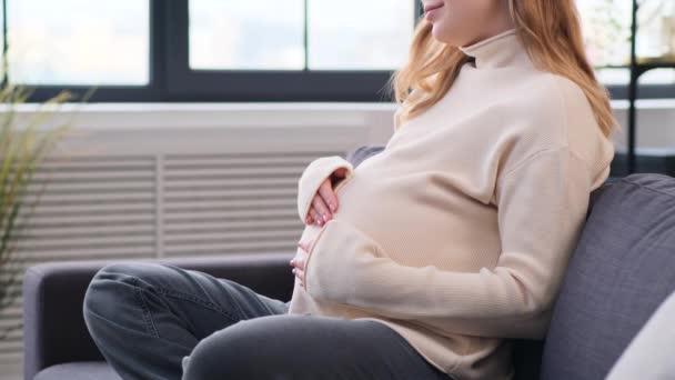 Close-up tiro de uma jovem mulher branca grávida acariciando barriga, relaxando no sofá em casa sala de estar. Gravidez, parto, futuro conceito de paternidade. - Filmagem, Vídeo