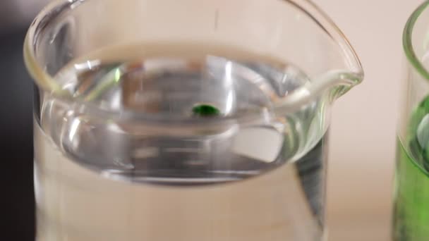 Primer plano de gotas líquidas verdes que caen en el agua desde la parte superior en la taza química de vidrio y se disuelven mientras se cambia el color de la mezcla. Experimente con fluidos en laboratorio con equipos modernos. - Metraje, vídeo