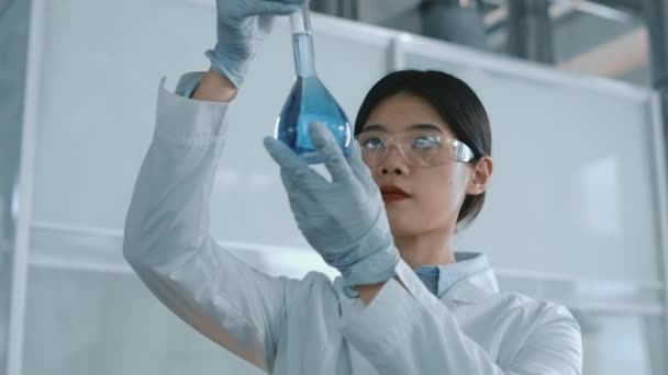 Střední detailní záběry mladé čínské ženy v brýlích, bílém plášti a rukavicích, které drží baňku s světle modrou chemickou tekutinou, zkoumají konzistenci a rezidua, při práci na nové sloučenině - Záběry, video