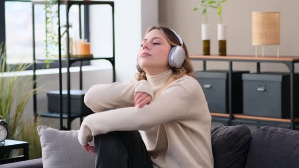 Mujer joven caucásica melancólica y sin esperanza escuchando música en los auriculares, sentada en el sofá en casa. Tristeza y apatía durante el fin de semana. - Imágenes, Vídeo