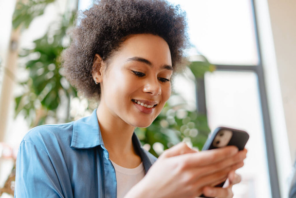 Młoda, atrakcyjna Afroamerykanka z kręconymi włosami, używająca telefonu komórkowego, SMS-a, zbliżenie. Koncepcja technologii internetowych, reklama - Zdjęcie, obraz