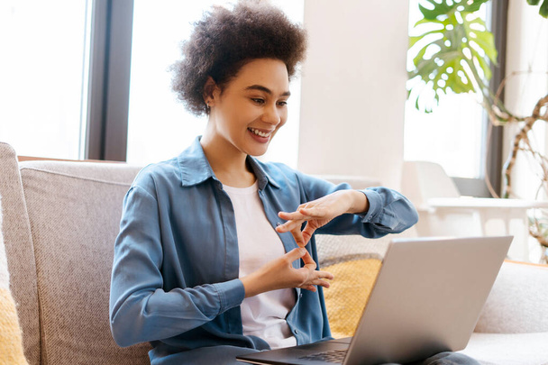 Счастливая веселая афроамериканка, пользующаяся ноутбуком, имеющая видеоконференцию, общающаяся с жестовым языком, сидящая дома на удобном диване. Концепция коммуникации - Фото, изображение