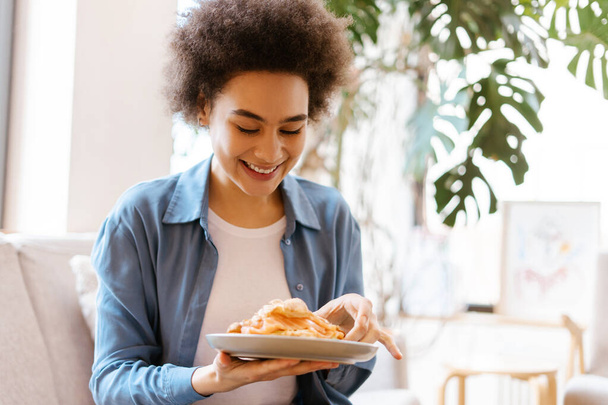Beautiful, smiling African American woman holding croissant while sitting on comfortable sofa in room. Concept of break, snack, meal, morning routine - Photo, Image