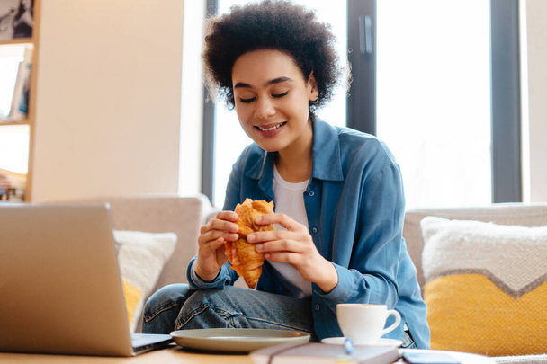 Femme afro-américaine élégante et souriante aux cheveux bouclés, indépendante assise sur un canapé confortable à l'aide d'un ordinateur portable, tenant un croissant frais, faisant une pause, grignotant dans un café. Concept de nourriture, déjeuner, travail à distance - Photo, image