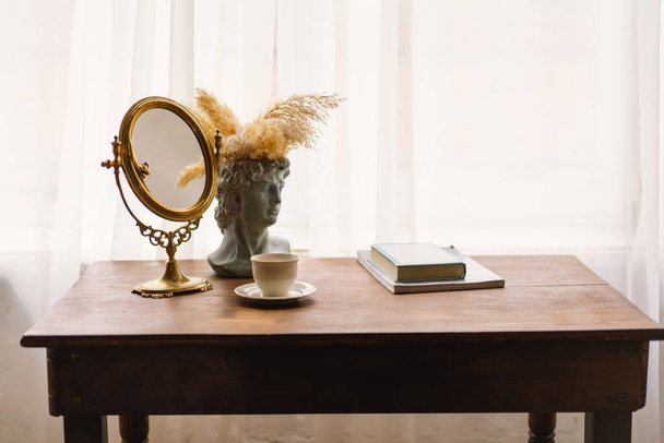 Espelho moldado a ouro fica em uma mesa de madeira, ao lado de um vaso de busto clássico adornado com grama pampas. Uma simples xícara de café branco completa o ambiente calmante e sofisticado. - Foto, Imagem