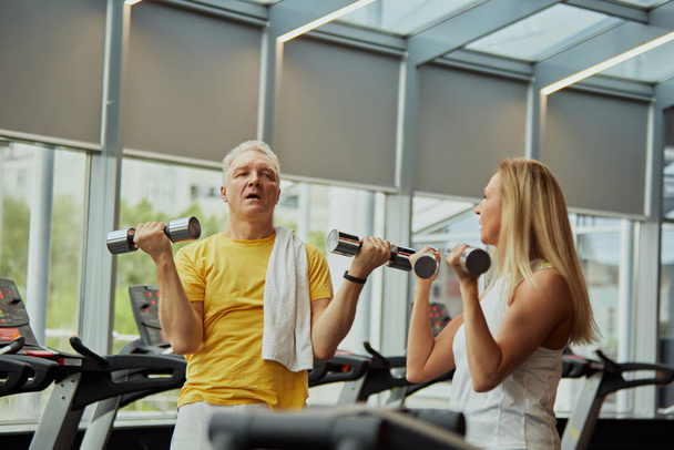 Ώριμο ζευγάρι, άνδρας και γυναίκα στα αθλητικά κάνοντας ασκήσεις για τα χέρια με αλτήρες στο σύγχρονο γυμναστήριο. Έννοια του αθλητισμού, ενεργό ηλικιωμένους στη σύγχρονη ζωή, υγιεινό τρόπο ζωής, γυμναστήριο για συνταξιοδοτήσεις. - Φωτογραφία, εικόνα