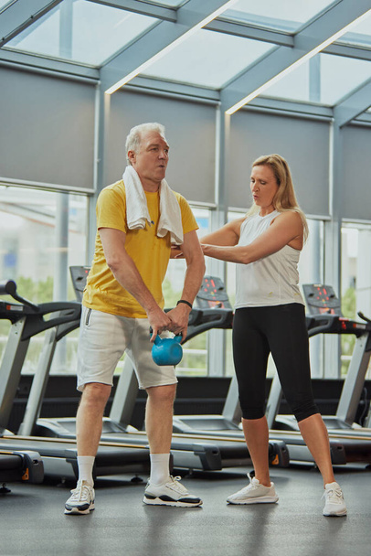Oudere man doet oefeningen, tilt gewichten en vrouw verzekert hem in een modern fitnesscentrum voor gepensioneerden. Concept van sport, actieve senioren in het moderne leven, gezonde levensstijl. Steun - Foto, afbeelding