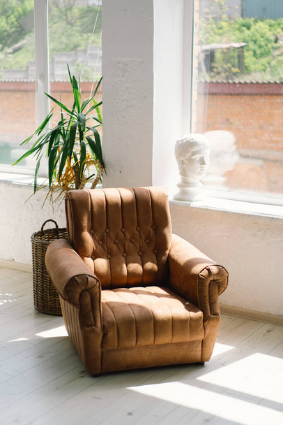 A vintage leather armchair basks in sunlight near a large window with a clear view outside, accompanied by a green potted plant and a wicker basket in a serene, bright room. - Photo, Image