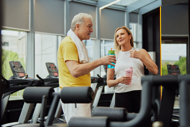 El dúo de fitness para ancianos comparte conversación e hidratación después del entrenamiento en el gimnasio con equipos modernos. Concepto de deporte, personas mayores activas en la vida moderna, estilo de vida saludable. Anuncio - Foto, imagen