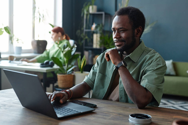 Portrait of smiling African American man using laptop at workplace in open office decorated with green plants copy space - Photo, Image