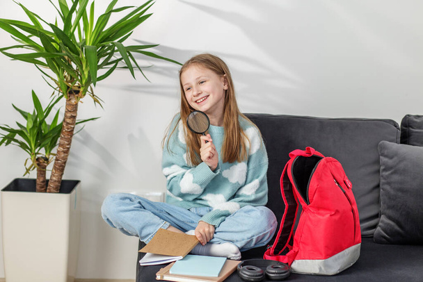 Usmívající se mladá dívka sedí se zkříženýma nohama na pohovce drží lupu, se školní sešity a pulzující červený batoh vedle ní. - Fotografie, Obrázek