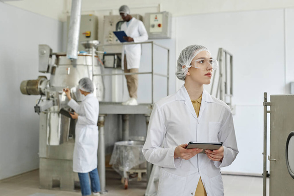 Retrato de cintura hacia arriba de una mujer joven con bata de laboratorio y tableta en taller limpio de fábrica farmacéutica, espacio para copiar - Foto, Imagen