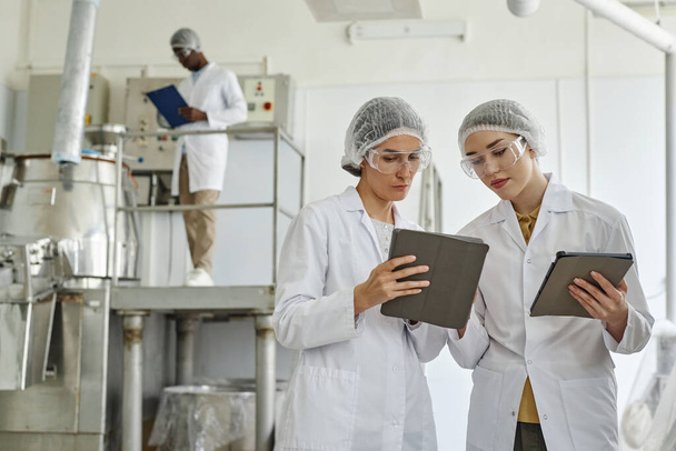 Objímání portrétu dvou mladých žen v laboratorních pláštích a použití digitálního tabletu v dílně farmaceutické továrny, kopírovací prostor - Fotografie, Obrázek