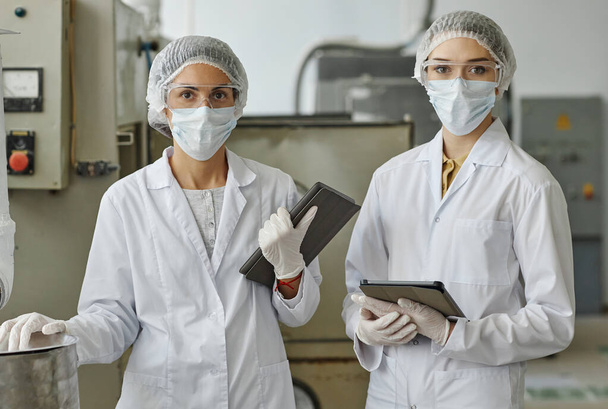 Retrato de cintura hacia arriba de dos mujeres jóvenes con máscaras y batas de laboratorio mirando a la cámara mientras trabajan en fábrica - Foto, Imagen