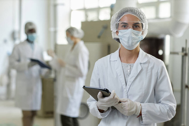Retrato de cintura hacia arriba de una mujer joven con máscara y bata de laboratorio mirando a la cámara en el taller de fábrica, espacio para copiar - Foto, Imagen