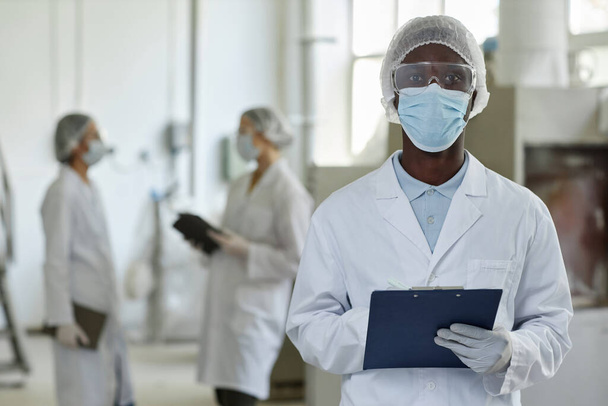 Maskeli ve laboratuvar önlüklü genç bir adamın belden yukarı portresi. Fabrika atölyesinde kameraya bakıyor. - Fotoğraf, Görsel